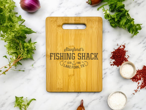 A personalized bamboo cutting board with a charming fishing shack illustration. The board is ideal for kitchen use and features a name or initials.